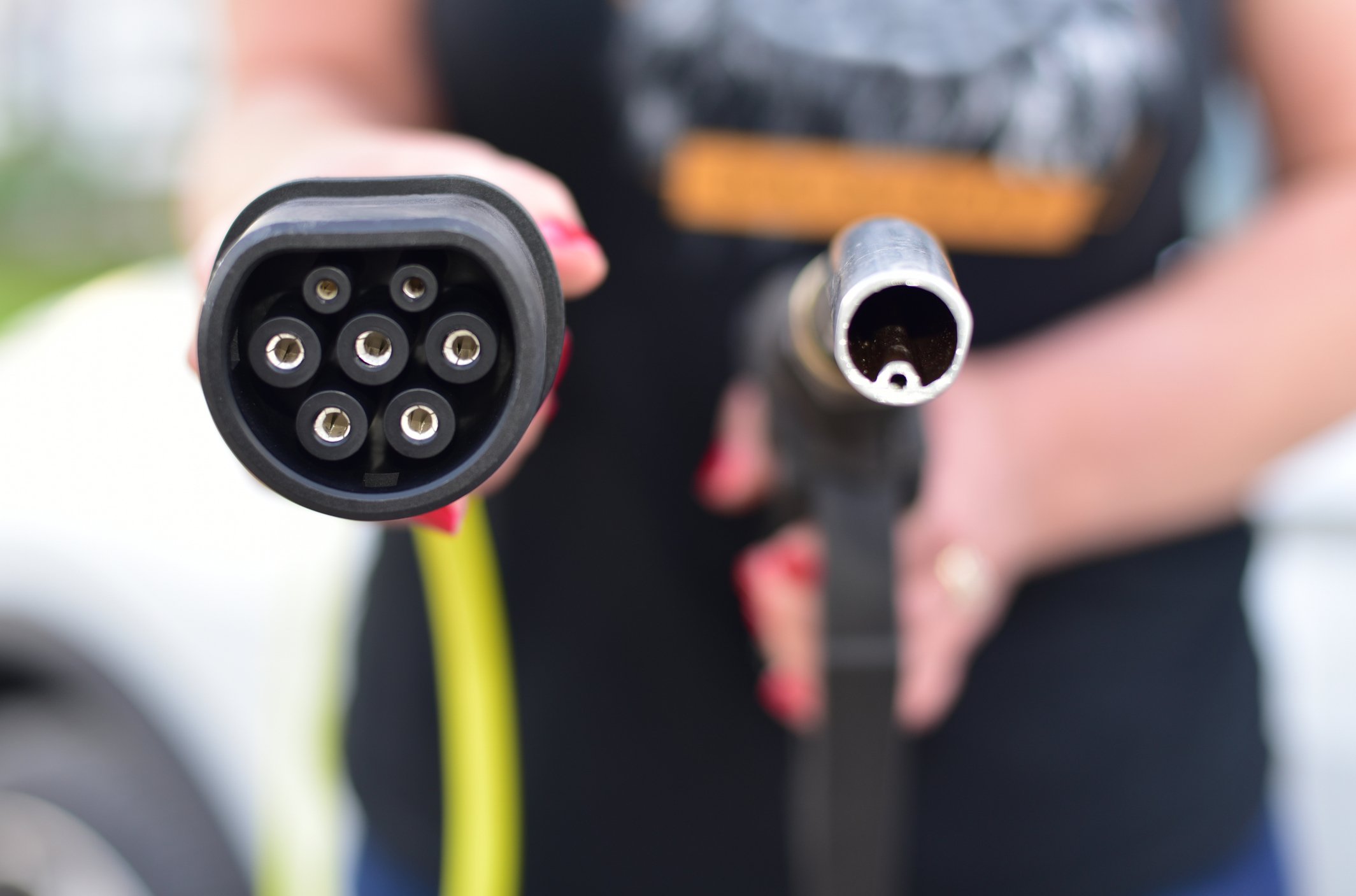 Personal holding a gasoline pump in one hand and an electronic vehicle charging plug in the other. 